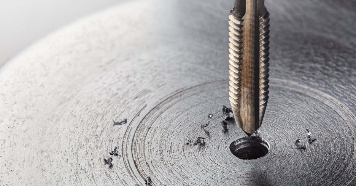 A thread tap drill bit near a piece of metal. A hole lies underneath the drill bit with small pieces of material.